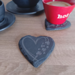 Heart-shaped engraved slate coaster with a delicate heart and shamrock design, placed on a wooden surface with a coffee cup in the background.
