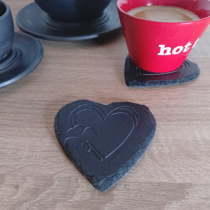 A heart-shaped engraved slate coaster placed on a wooden surface, with a red coffee cup resting on another coaster in the background.
