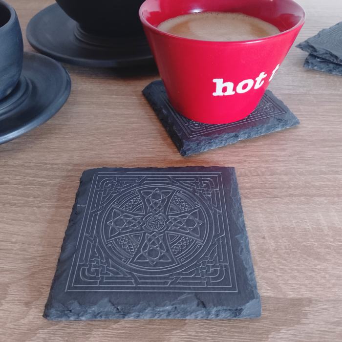 A square engraved slate coaster placed on a wooden surface, with a red coffee cup resting on another coaster in the background.