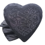 A heart-shaped slate coaster with a detailed Celtic dragon engraving, stacked against other coasters.