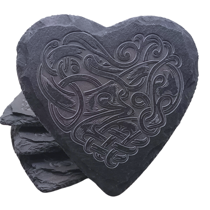 A heart-shaped slate coaster with a detailed Celtic dragon engraving, stacked against other coasters.