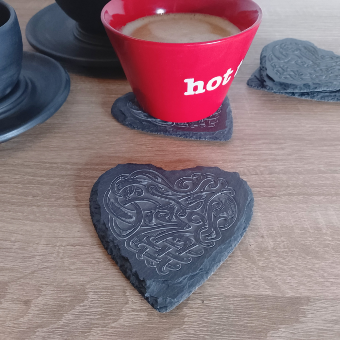 A heart-shaped engraved slate coaster placed on a wooden surface, with a red coffee cup resting on another coaster in the background.