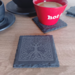 A square engraved slate coaster placed on a wooden surface, with a red coffee cup resting on another coaster in the background.