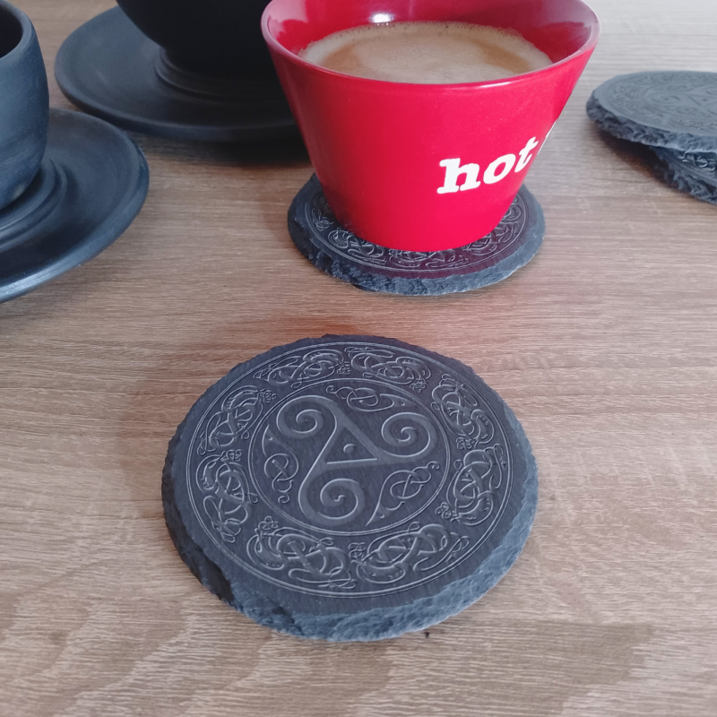 A round engraved slate coaster placed on a wooden surface, with a red coffee cup resting on another coaster in the background.