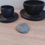 Engraved Triquetra stone placed on a wooden surface, accompanied by black ceramic cups, creating a calm and elegant setting.