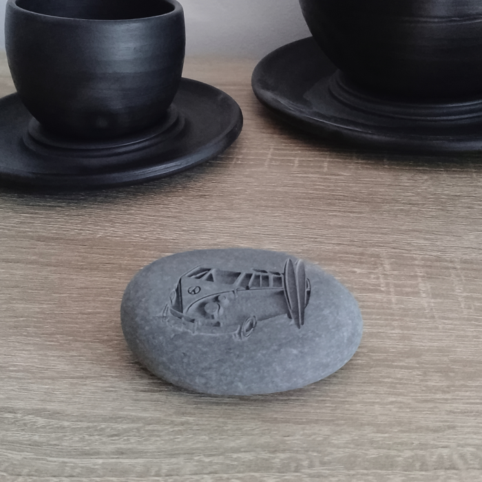 A natural engraved stone featuring a classic surf van and surfboards, placed on a wooden surface with black ceramic cups in the background.