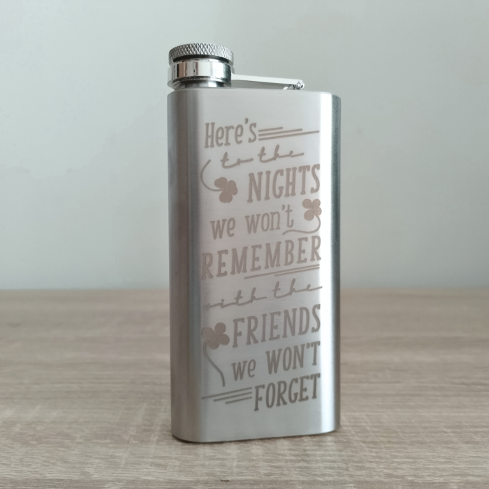 Silver stainless steel custom flask engraved with humorous Irish toast, upright on wooden table.